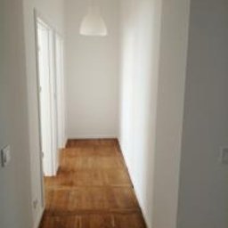 Katarzyna - Wąski, biały korytarz po remoncie z parkietem w jodełkę. Widoczne drzwi po lewej stronie i minimalistyczna lampa wisząca. Ściany świeżo pomalowane na biało.