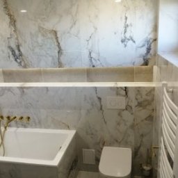 Katarzyna - Elegancka łazienka z marmurowymi płytkami, złotą armaturą i grzejnikiem drabinkowym. Umywalka wpuszczana w blat, minimalistyczna toaleta podwieszana.