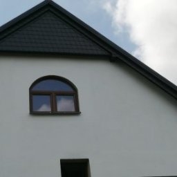 Katarzyna - Elewacja domu w Wołowie z oknem w kształcie półkola i ciemnym dachem. Widok na szczyt budynku z bliska, jasna ściana i fragment błękitnego nieba.