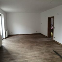 Katarzyna - Jasne wnętrze po remoncie: drewnopodobna podłoga, świeżo pomalowane białe ściany, grzejniki, widoczne gniazdka elektryczne i puszki instalacyjne. Minimalizm.