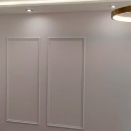 Katarzyna - Minimalistyczne wnętrze: białe ściany z dekoracyjnymi ramami, złote wiszące lampy z okrągłymi kloszami i sufitowe lampy w kształcie pierścieni.