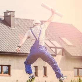 Entuzjastyczny budowlaniec w kombinezonie i kasku skacze z radości przed domem, trzymając zwój planów. Optymizm w budownictwie.