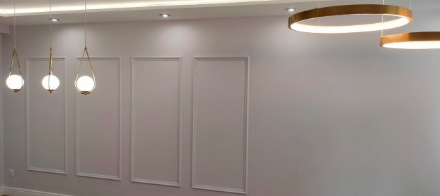 Minimalistyczne wnętrze: białe ściany z dekoracyjnymi ramami, złote wiszące lampy z okrągłymi kloszami i sufitowe lampy w kształcie pierścieni.