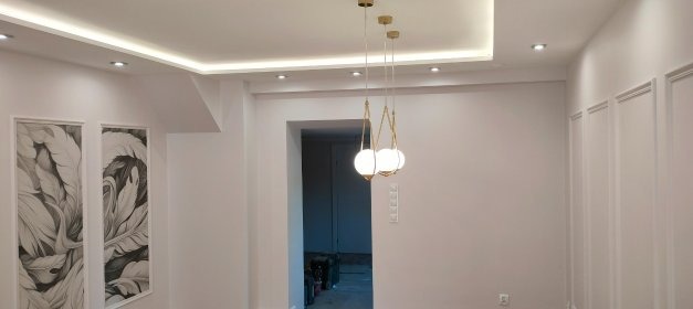 Białe wnętrze po remoncie: dekoracyjne panele ścienne, podwieszany sufit z oświetleniem LED, wisząca lampa i obrazy z motywem liści. Wykończenie w stylu nowoczesnym.