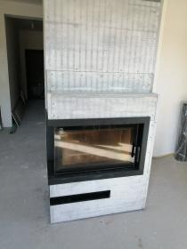 Betonowy kominek z czarną ramą w surowym wnętrzu. Widoczna struktura betonu i niedokończone wykończenie podłogi. Minimalistyczny design.