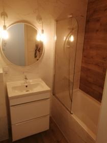 Jasna łazienka po remoncie: biała szafka z umywalką, okrągłe lustro z oświetleniem, przeszklony parawan nawannowy, imitacja drewna na ścianie.