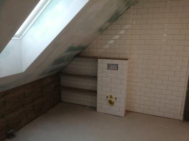 Łazienka na poddaszu po remoncie: białe, prostokątne płytki na ścianach, drewnopodobna okładzina i wnęki na półki. Skos z oknem dachowym. Minimalistyczny styl.