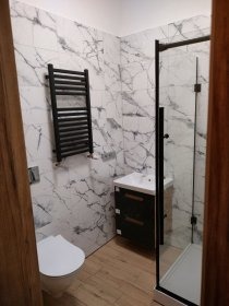 Elegancka łazienka w bieli i czerni: kabina prysznicowa z czarną ramą, marmurowe płytki ścienne, drewnopodobna podłoga i czarny grzejnik drabinkowy.