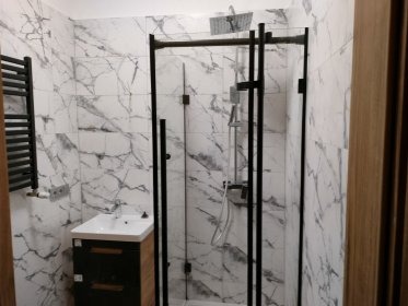 Nowoczesna łazienka w bieli i czerni: marmurowe płytki, kabina prysznicowa typu walk-in z czarnymi profilami, szafka umywalkowa z drewnianym akcentem.