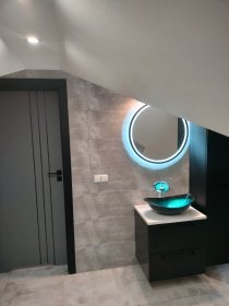 Nowoczesna łazienka na poddaszu: szara ściana, umywalka z turkusowego szkła i okrągłe lustro z podświetleniem LED, czarne szafki i szare drzwi.