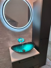 Turkusowa, owalna umywalka na szafce z blatem, okrągłe lustro z podświetleniem LED i szara ściana w nowoczesnej łazience. Nietypowy design wnętrza.