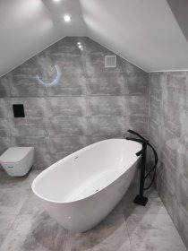 Nowoczesna łazienka na poddaszu: biała wanna wolnostojąca, szare płytki imitujące marmur, czarna armatura i podwieszana toaleta. Minimalistyczny design z oświetleniem LED.