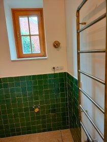 Łazienka z zielonymi kaflami do połowy ściany, drewniane okno, grzejnik drabinkowy i mosiężna armatura. Nietypowe połączenie kolorów i materiałów.