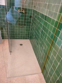 Wykończona kabina prysznicowa z zielonymi, kwadratowymi płytkami na ścianach i jasnym brodzikiem. Widoczna szklana ściana kabiny i złota armatura.