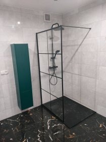 Nowoczesna kabina prysznicowa z czarnymi profilami na tle marmurowej podłogi i białych płytek. Ciemnozielona szafka łazienkowa w tle.