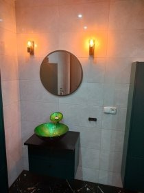 Wykończona łazienka w Wołowie: szare płytki, okrągłe lustro, zielona umywalka na czarnej szafce, oświetlenie ścienne. Nowoczesny design, ciepłe światło.