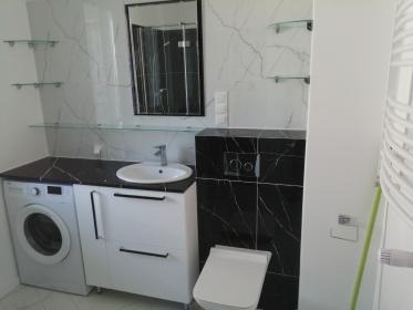 Elegancka łazienka z marmurowymi płytkami, czarnym blatem i szafką pod umywalką. Widoczna pralka, lustro w czarnej ramie i szklane półki. Nowoczesny design.