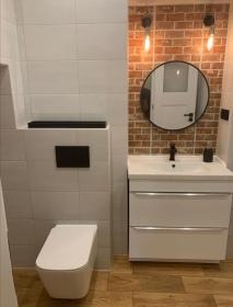 Nowoczesna łazienka po remoncie: biała umywalka z szafką, lustro w czarnej ramie na tle ściany z cegły i podwieszana toaleta. Drewnopodobna podłoga.
