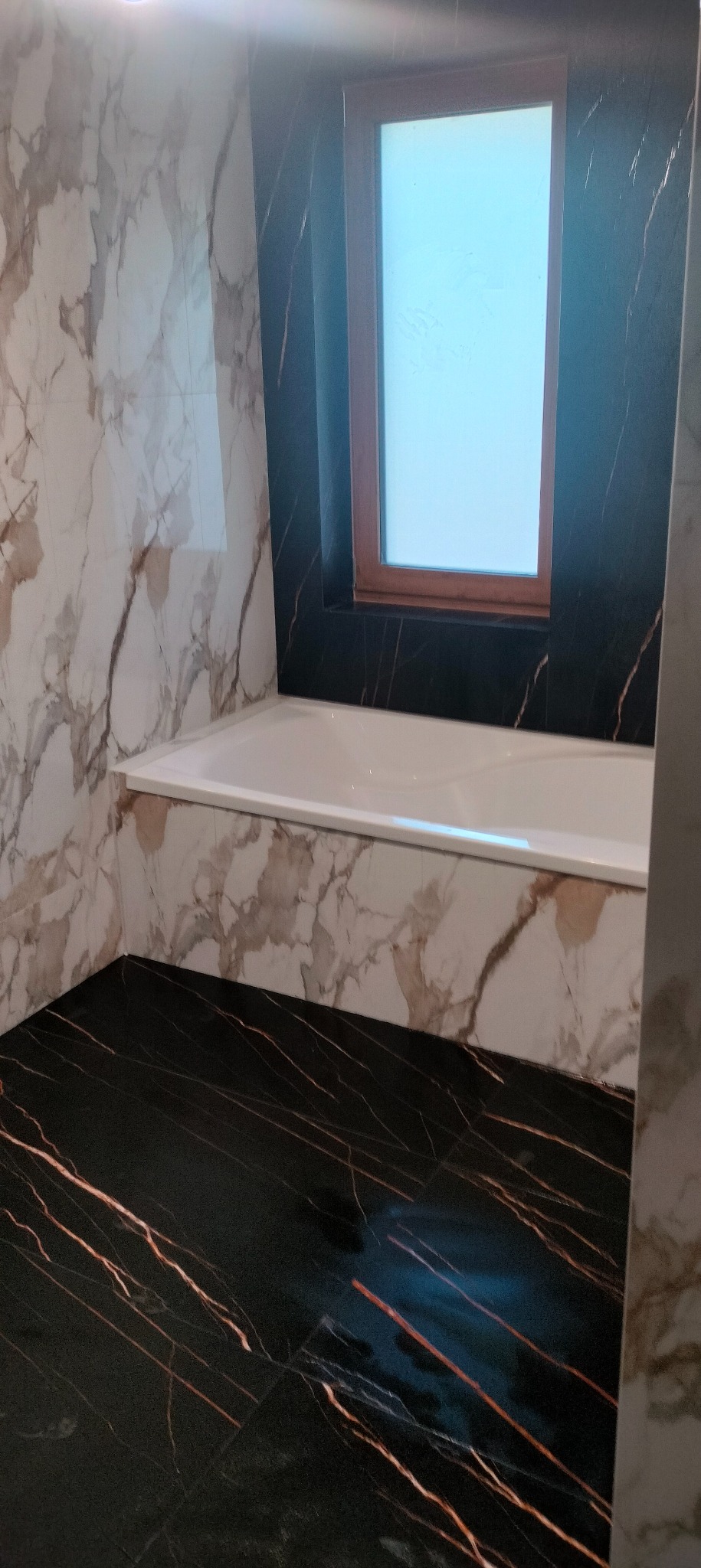 Wykończona łazienka z wanną, marmurowymi płytkami na ścianach i czarną podłogą z żyłkami. Okno w drewnianej ramie nad wanną. Nowoczesny design.