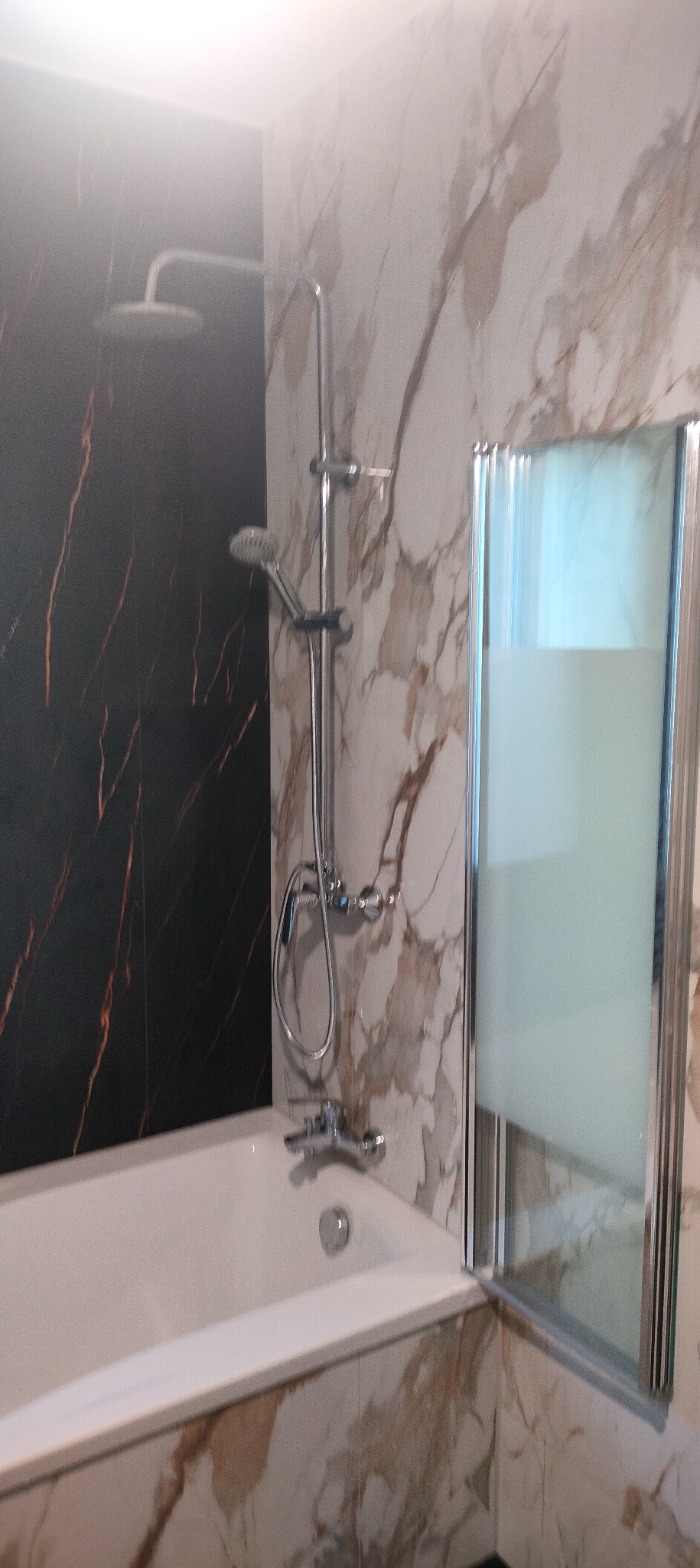 Wykończona łazienka z wanną, chromowaną armaturą i panelami imitującymi marmur w odcieniach bieli, beżu i czerni. Kabina prysznicowa z matową szybą.