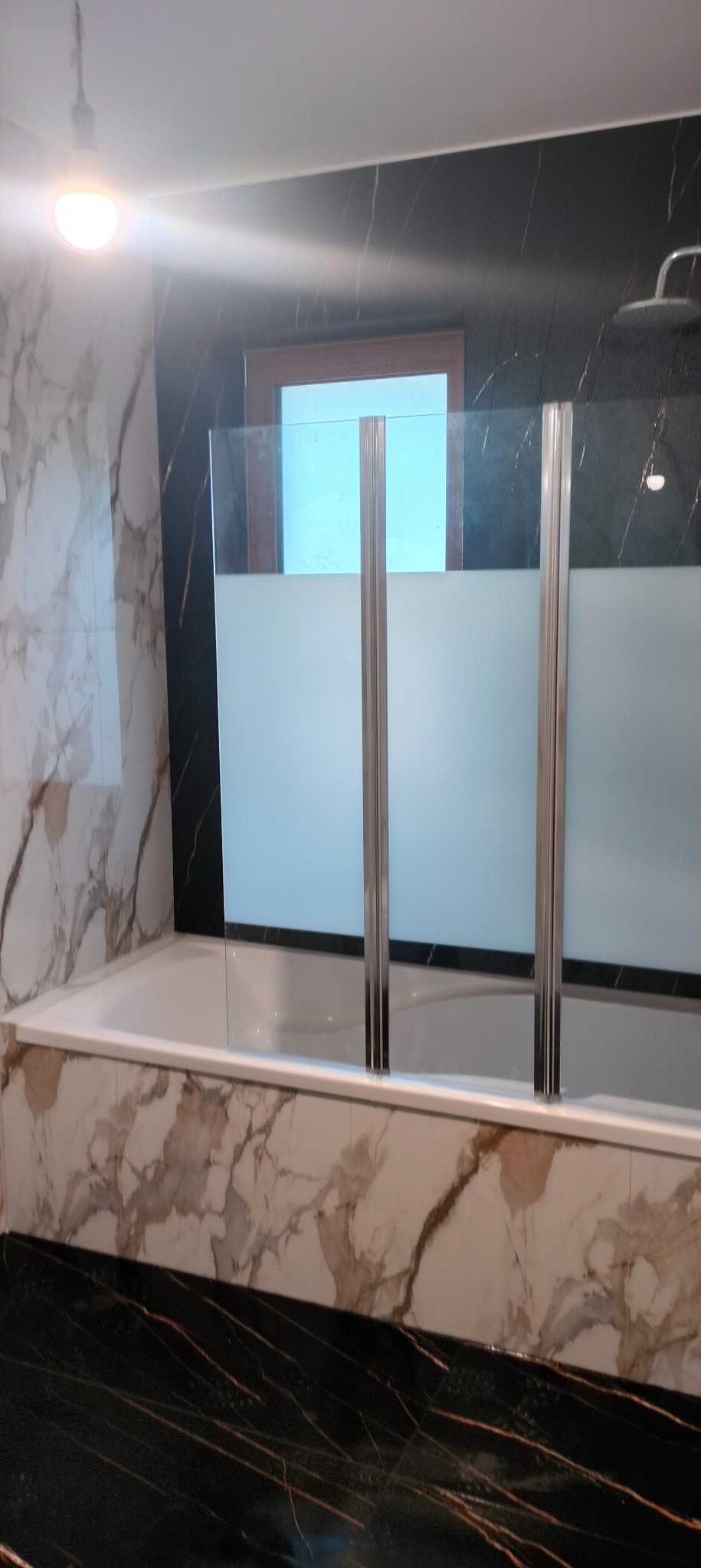 Biała wanna z marmurową obudową i szklanym parawanem z chromowanymi profilami na tle ściany z czarnego marmuru z białymi żyłami. Widoczny fragment sufitu i prysznica.