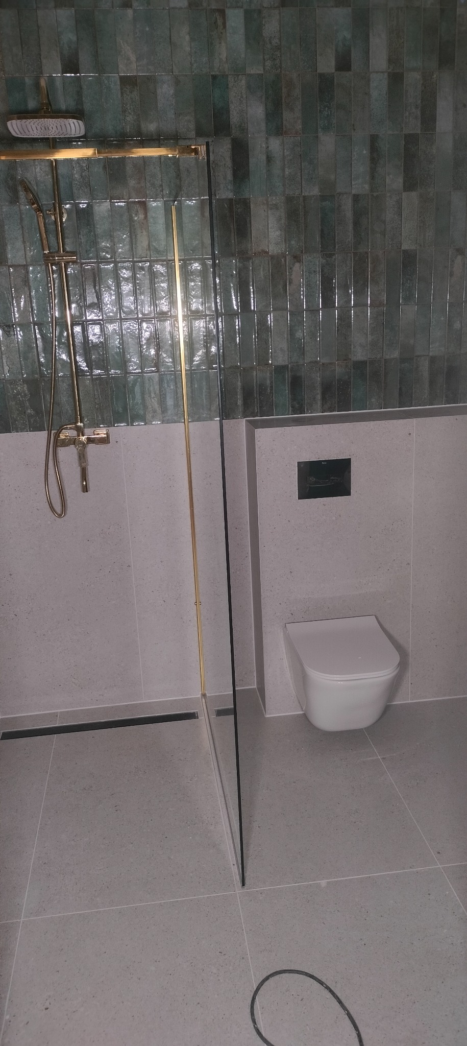 Nowoczesna łazienka z kabiną prysznicową typu walk-in, zielonymi płytkami na ścianie i podwieszaną toaletą. Minimalistyczny design z elementami w kolorze złotym.