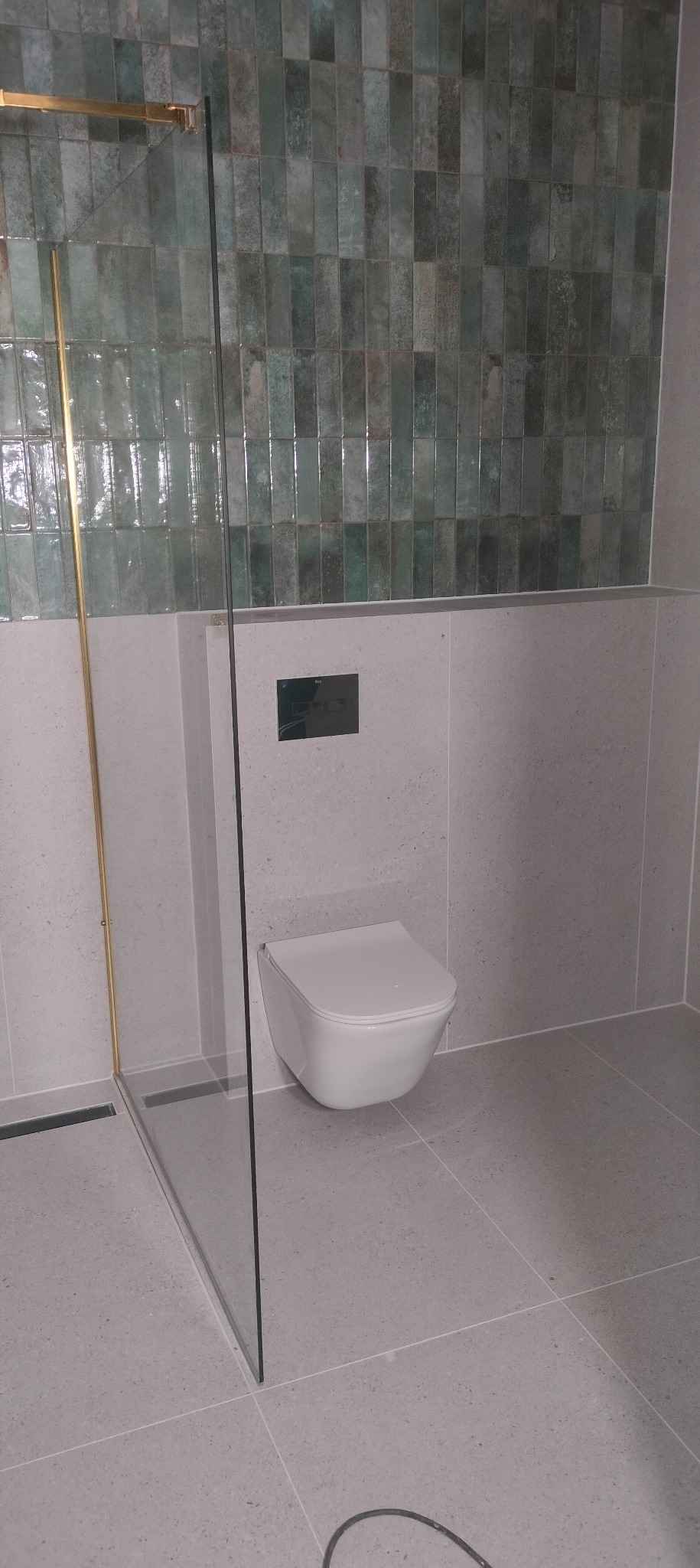 Nowoczesna łazienka z kabiną prysznicową ze złotymi detalami, wiszącą toaletą i zielonymi, błyszczącymi płytkami na ścianie. Minimalistyczny design.