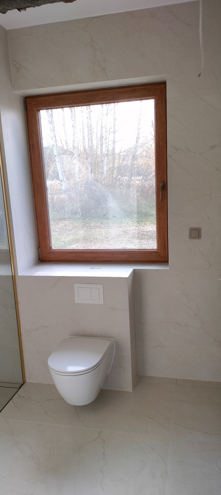 Nowoczesna łazienka z oknem w drewnianej ramie. Biała ceramika, płytki imitujące marmur. Widok na drzewa za oknem. Minimalistyczny design.