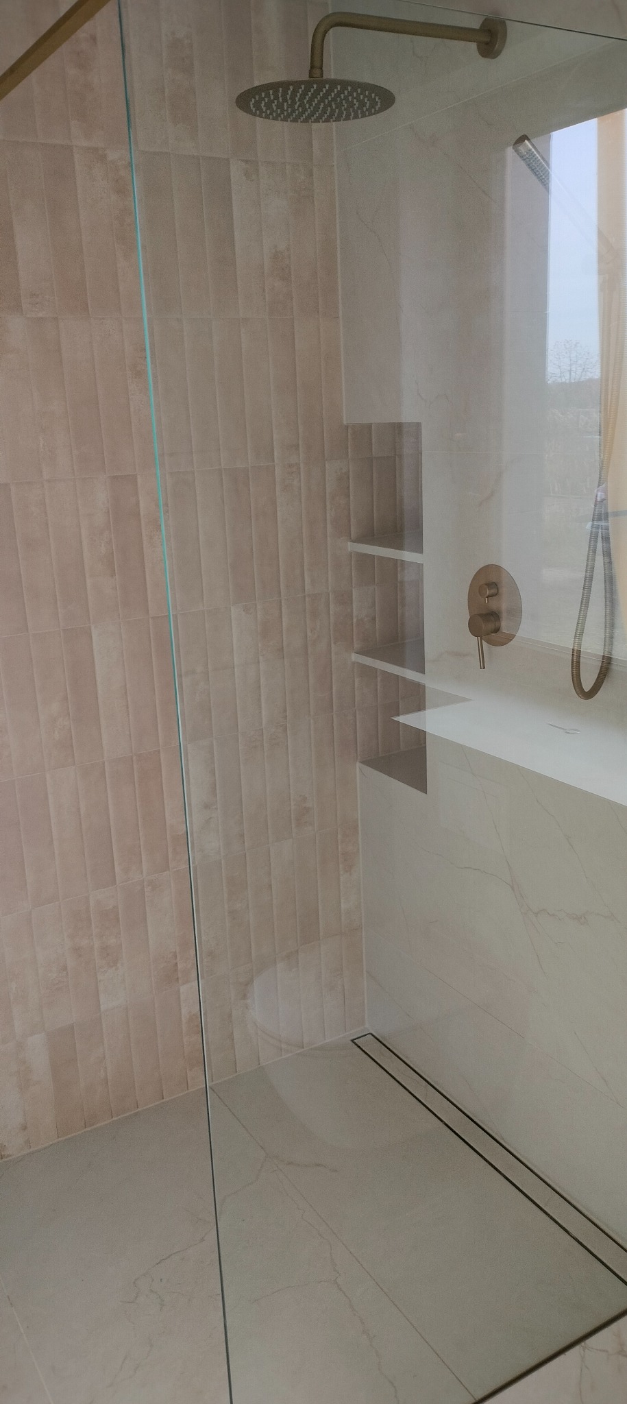 Beżowa kabina prysznicowa walk-in z odpływem liniowym, armaturą w kolorze starego złota i wnęką z półkami. Minimalistyczny design, jasne płytki.