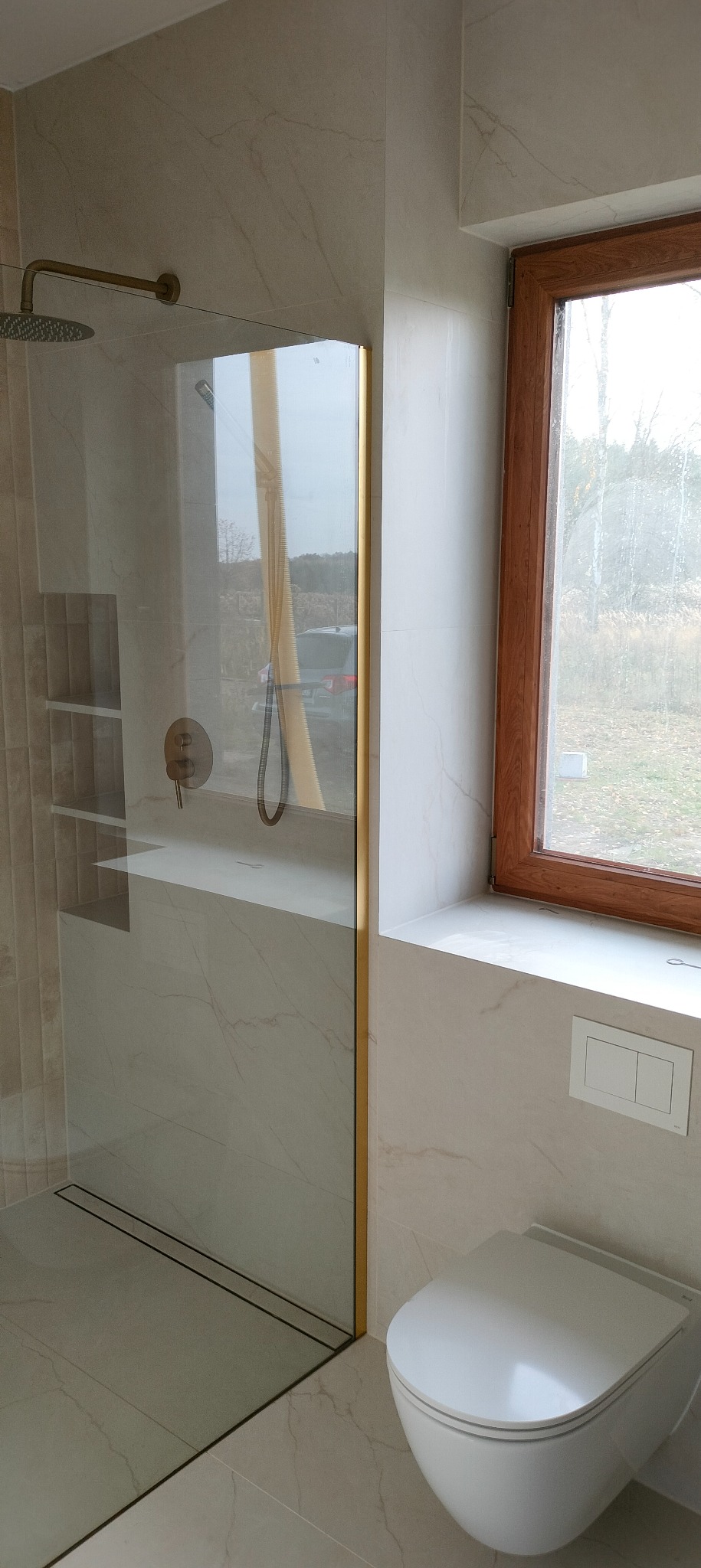 Beżowa łazienka z kabiną prysznicową ze złotymi detalami, widok na okno i podwieszaną toaletę. Minimalistyczny design z marmurowymi płytkami.