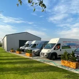 Flota firmowa: kilka vanów z logo fotowoltaiki, klimatyzacji i pomp ciepła zaparkowanych przed budynkiem magazynowym na zadbanym trawniku.