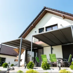 Nowoczesna pergola z szarą tkaniną zacieniającą, przylegająca do białego domu z brązowym dachem. Widoczne zielone krzesła i zadbana roślinność.