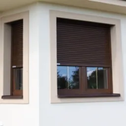 Brązowe rolety zewnętrzne na dwóch oknach budynku z jasną elewacją i beżowymi obramowaniami okiennymi, widok z zewnątrz.