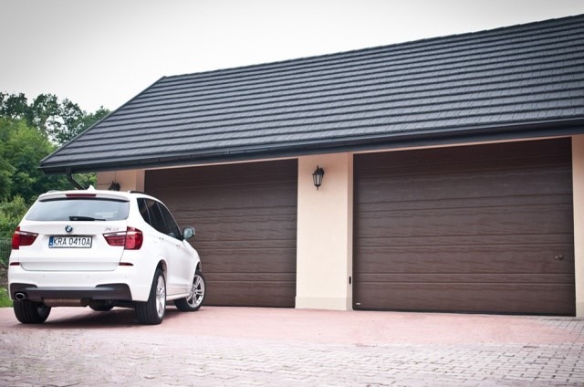 Dwustanowiskowy garaż z brązowymi bramami segmentowymi, przed którym stoi białe BMW X3. Dach pokryty grafitową dachówką.