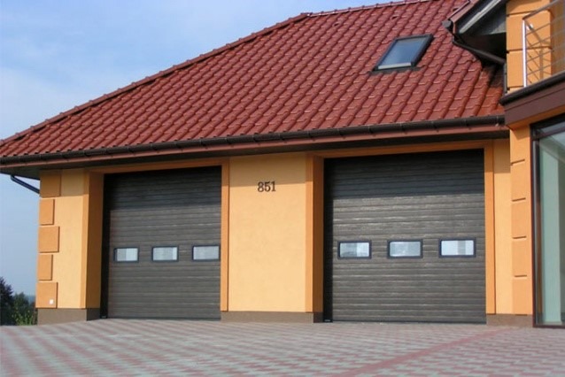 Dwie brązowe bramy garażowe segmentowe z oknami, w budynku z pomarańczową elewacją i czerwoną dachówką. Nad bramami widoczny numer domu '851'.