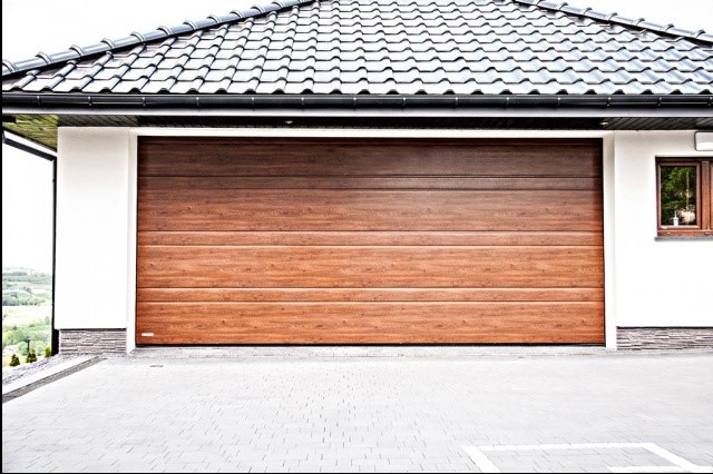Nowoczesna brama garażowa segmentowa w kolorze drewna, z poziomymi panelami, zamontowana w białym garażu z ciemnym dachem. Widok z zewnątrz, na tle kostki brukowej.