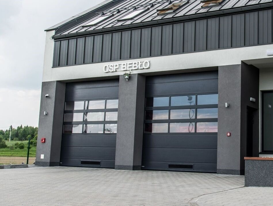 Dwie grafitowe bramy garażowe z przeszkleniami w budynku OSP Bebło, widoczne z zewnątrz. Ujęcie z poziomu chodnika, w tle zieleń.