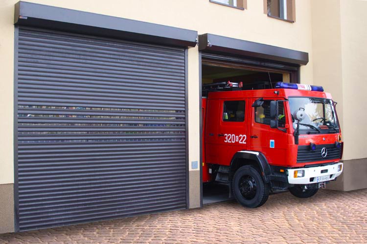 Ciemna brama garażowa segmentowa z poziomymi przetłoczeniami na tle budynku straży pożarnej, z wjazdu wyjeżdża czerwony wóz strażacki.