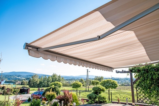 Zewnętrzna markiza tarasowa w beżowe paski, rozłożona nad patio z widokiem na krajobraz. Falbana na krawędzi, szara konstrukcja, błękitne niebo w tle.