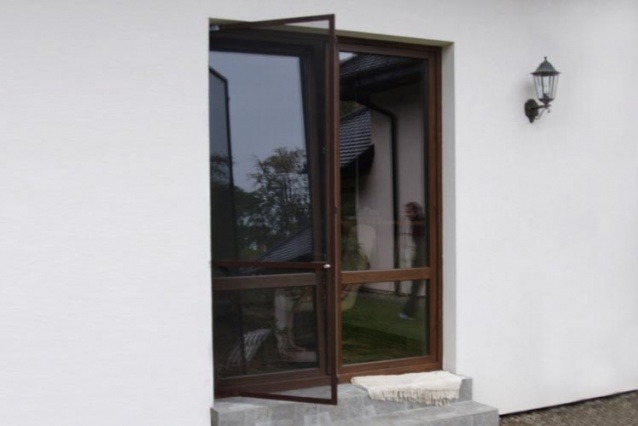 Brązowe drzwi balkonowe otwarte na zewnątrz, z widocznym odbiciem otoczenia w szybach. Biała ściana budynku i lampa zewnętrzna. Wejście na schodach.
