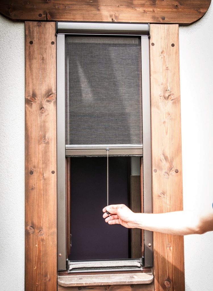 Zewnętrzna roleta okienna z drewnianą ramą, częściowo opuszczona, obsługiwana ręcznie. Widoczna faktura ściany i fragment ręki. Ujęcie z bliska.