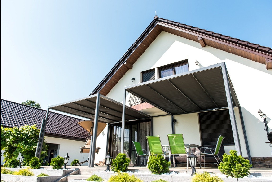 Nowoczesna pergola z szarą tkaniną zacieniającą, przylegająca do białego domu z brązowym dachem. Widoczne zielone krzesła i zadbana roślinność.