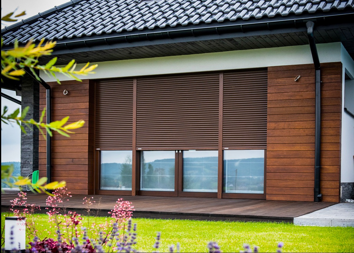 Elewacja domu z brązowymi roletami zewnętrznymi, częściowo zasłoniętymi. Widoczny fragment tarasu i zadbany trawnik z kwiatami. Ujęcie z dołu, podkreślające nowoczesny design.