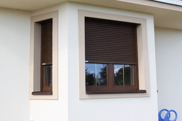 Brązowe rolety zewnętrzne na dwóch oknach budynku z jasną elewacją i beżowymi obramowaniami okiennymi, widok z zewnątrz.