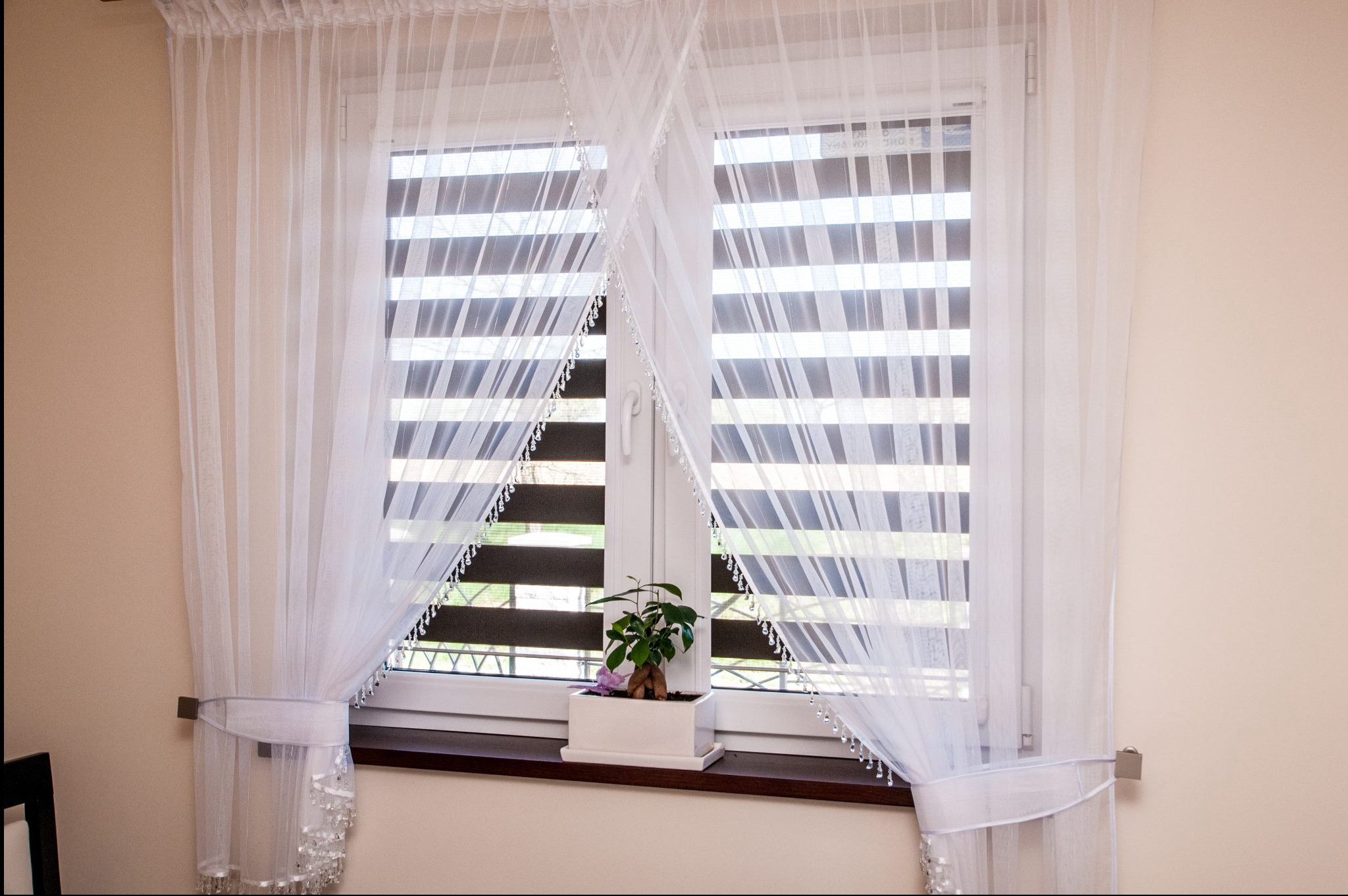Okno z roletami dzień noc, udekorowane zwiewnymi, białymi firanami z ozdobnymi koralikami. Na parapecie stoi doniczka z zieloną rośliną.