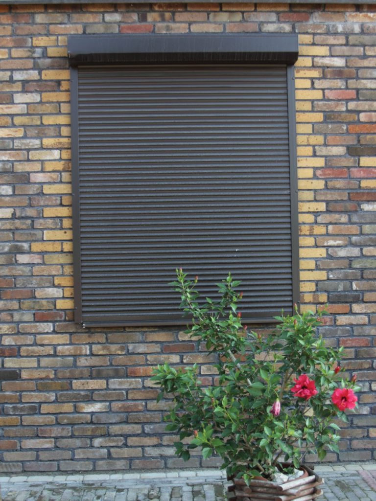 Zasłonięta brązowa roleta zewnętrzna na tle ceglanej ściany budynku, z doniczką z kwitnącym hibiskusem u dołu kadru. Widok z zewnątrz.