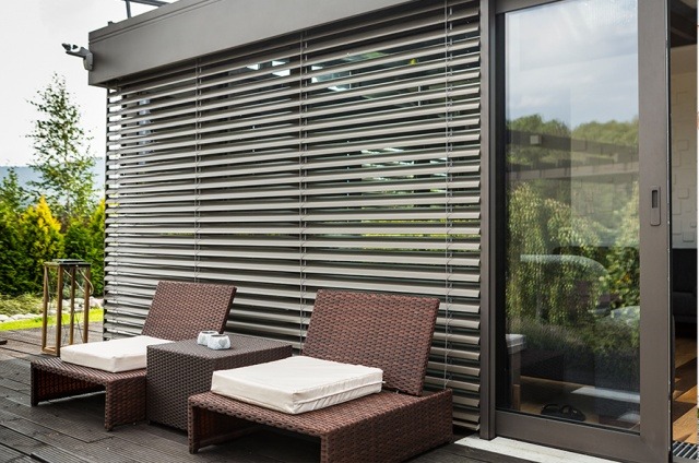 Zewnętrzne, poziome rolety w kolorze szarym, częściowo zasłonięte, zamontowane na tarasie z brązowymi meblami ogrodowymi i widokiem na zieleń.
