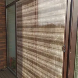 Zewnętrzna roleta plisowana w brązowej ramie, zamontowana na oknie budynku z cegły. Widoczny fragment drewnianego tarasu.