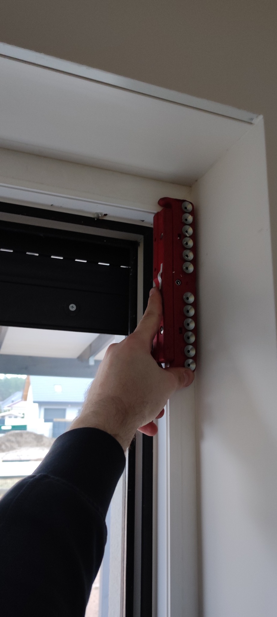 Pomiar okna roletą zewnętrzną z użyciem czerwonego narzędzia pomiarowego, widok z bliska na profil okna i dłoń. Dom w tle, światło dzienne.