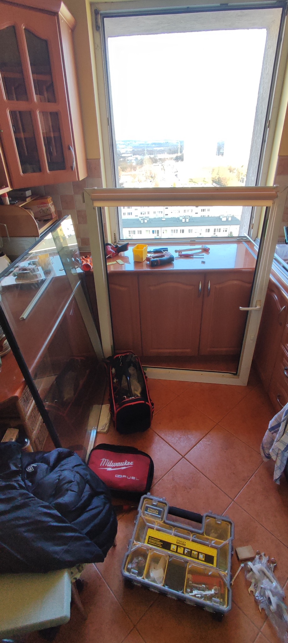 Demontaż okna balkonowego z widocznymi narzędziami i torbą narzędziową w pomieszczeniu z brązowymi płytkami podłogowymi i meblami.